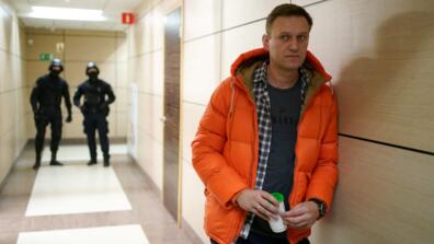 Alexei Navalny wears an orange jacket and leans against the wall of a business center which law enforcement officials watch from the background.