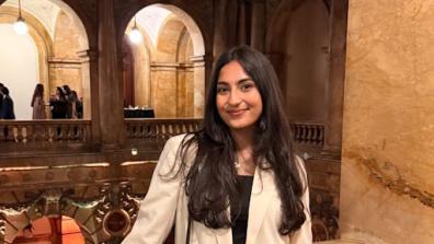 Photo of Farah  in a beige jacket with a black bag leaning against a railing  with tan archways in the background. 