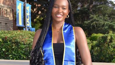 A photo of Samai graduating from her undergraduate institution. She is outside wearing a black dress with a blue graduation sash and green trees in the background. She is also holding her graduation cap.  