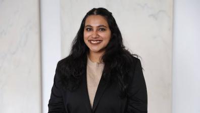 Headshot of Vanita wearing a tan top and black jacket with a beige background. 