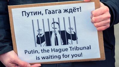 A bilingual poster showing Vladimir Putin behind bars and saying "Путин, Гаага ждёт!" (meaning "Putin, The Hague is waiting!") in Russian and "Putin, the Hague Tribunal is waiting for you!" in English
