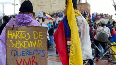Two protestors among others, one carrying a Colombian flag and the other one wearing a sign that says "Fed up with surviving, we want to live" in Spanish.