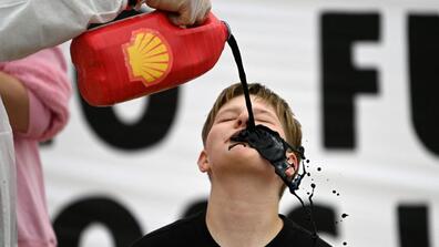 A child being poured crude oil to his mouth from a small gallon that has the logo of the oil company, Shell on it