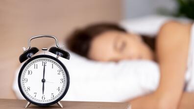 Close up of black alarm clock stand on bedside table show early morning hour, calm peaceful young woman sleep on background relax on fluffy pillow covered with warm blanket in bedroom