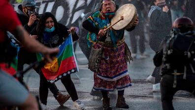 Protesters in the street. One is holding a colorful flag and another is dressed in traditional clothing and banging a drum.