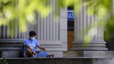 A college student wearing a mask sits in front of a building and works on their laptop