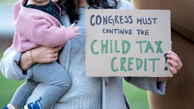 A woman carrying a baby with one arm and a banner saying "Congress must continue the child tax credit!" with the other