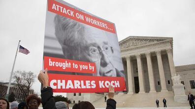 A protester holds up a sign that reads "attack on workers, brought to you by Charles Koch"
