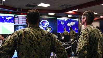 Two men in military uniform stand in front of a screen at a cyber command center.