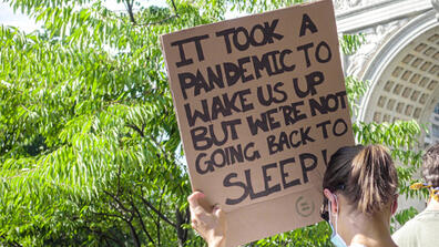 A person holds up a sign reading "it took a pandemic to wake us up, but we're not going back to sleep"