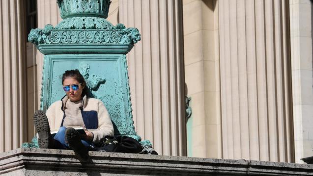 A student wearing sunglasses sits reading outside of Columbia's Low Library.