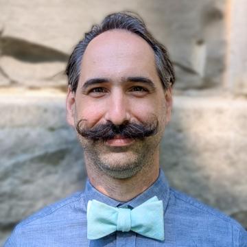 Headshot of Joseph Michael Lasky smiling in a blue shirt, light blue bow tie, and styled handlebar mustache.