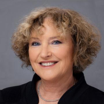 Headshot of a smiling Karnit Flug with blonde curly hair, wearing a black top and a thin necklace.