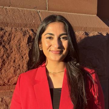 A photo of Farah smiling wearing a red jacket against a orange stone wall.