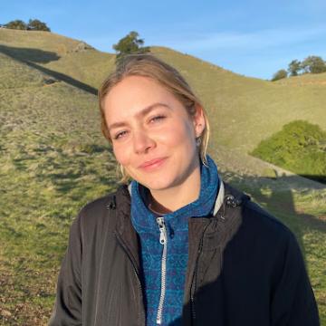 A photo of Hannah Taschbach outside wearing a blue sweater and black jacket with green hills behind her