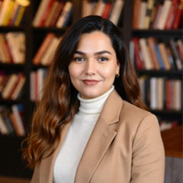 A pheadshot of Homaira wearing a tan blazer and a white turtleneck with a bookshelf in the background.