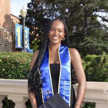 A photo of Samai wearing a black dress and a blue graduation sash. She is holding a graduation gap and is outside with trees behind her. 