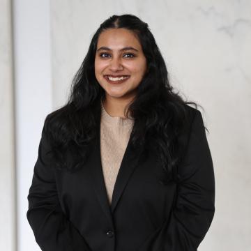 A photo of Vanita smiling wearing a black jacket with a cream wall in behind her. 