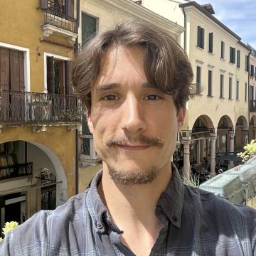 Headshot of Phd student Cameron Scalera smiling outdoors with European-style buildings in the background.
