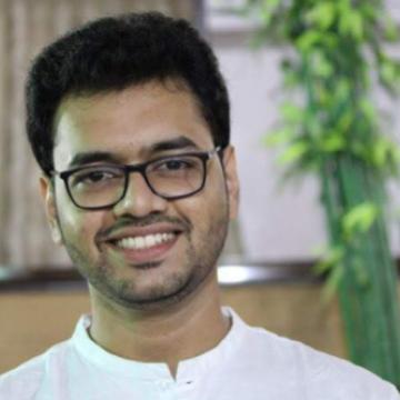 Headshot of Alhad wearing a white shirt and glasses with windows and greenery in the background.