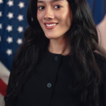 Headshot of Alya wearing a black top with the American flag in the background. 
