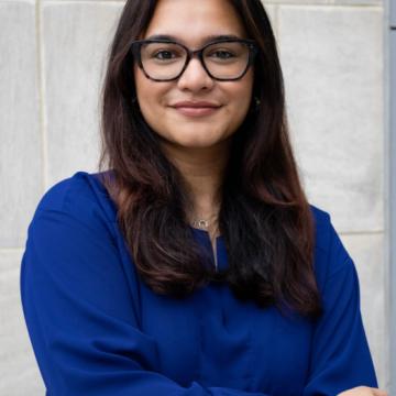 Headshot of Anushka wearing a blue top and glasses with a grey background.