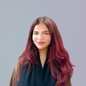 Headshot of Ashmi wearing a black top with red hair and a grey background. 