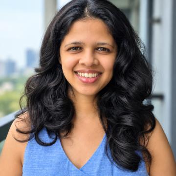 Headshot of Mugdha wearing a light blue top with the New York skyline in the background. 