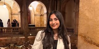 Photo of Farah  in a beige jacket with a black bag leaning against a railing  with tan archways in the background. 