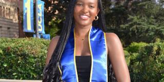 A photo of Samai graduating from her undergraduate institution. She is outside wearing a black dress with a blue graduation sash and green trees in the background. She is also holding her graduation cap.  