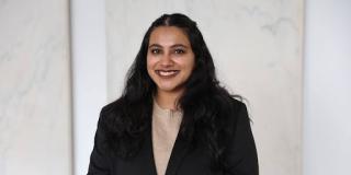 Headshot of Vanita wearing a tan top and black jacket with a beige background. 