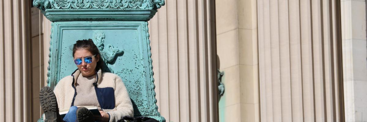 A student wearing sunglasses sits reading outside of Columbia's Low Library.