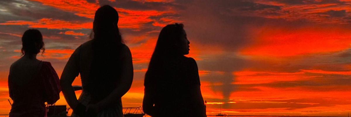 Silhouetted SIPA Capstone students observe a brilliant red-orange sunset over the water.